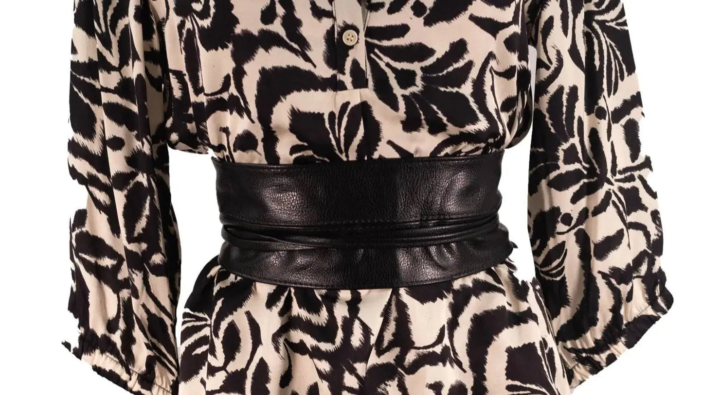 Cintura nera in pelle dal design avvolgente indossata sopra un abito con stampa astratta.