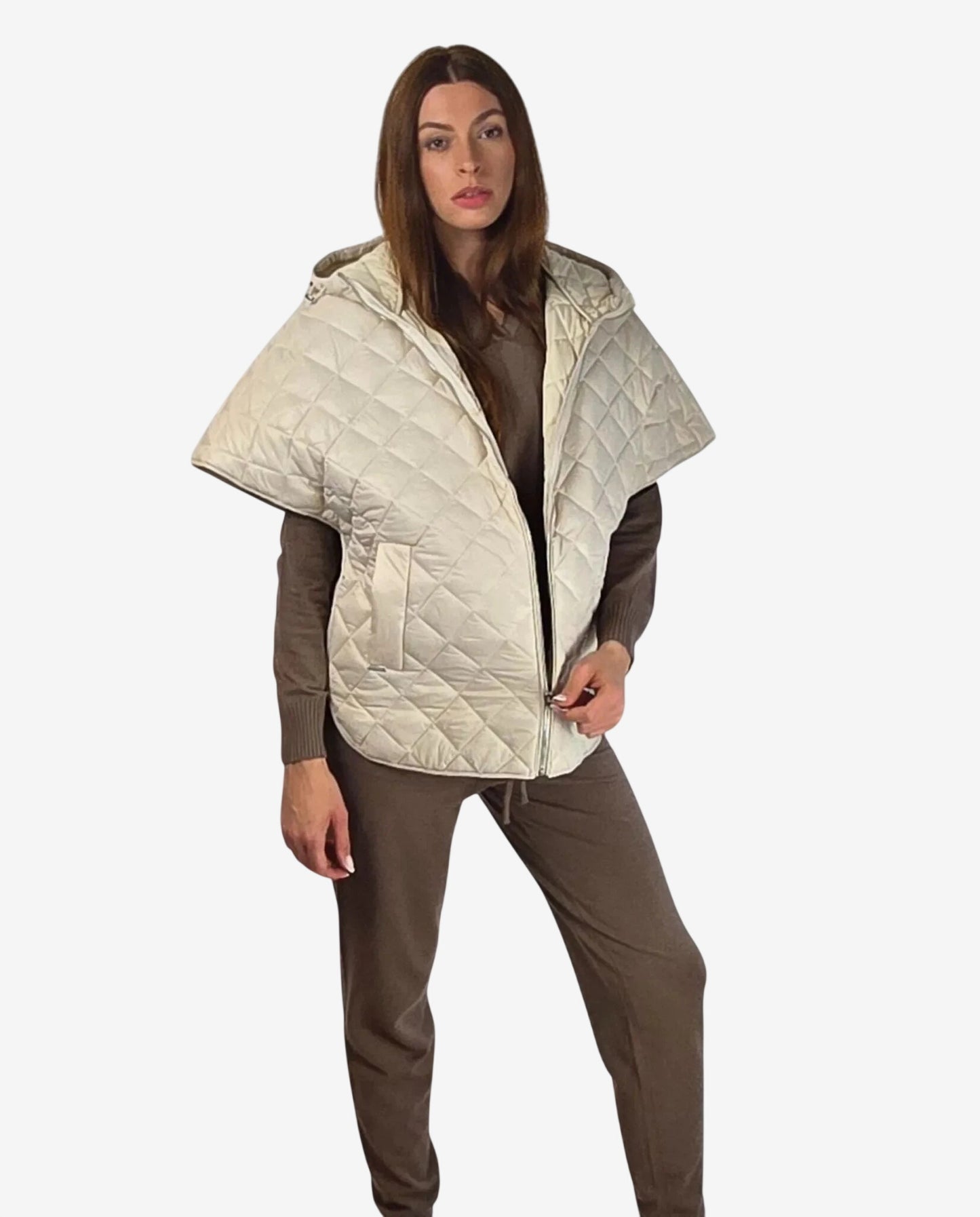 A woman wears a cream-colored, quilted hooded cape over a brown long-sleeved top and trousers.