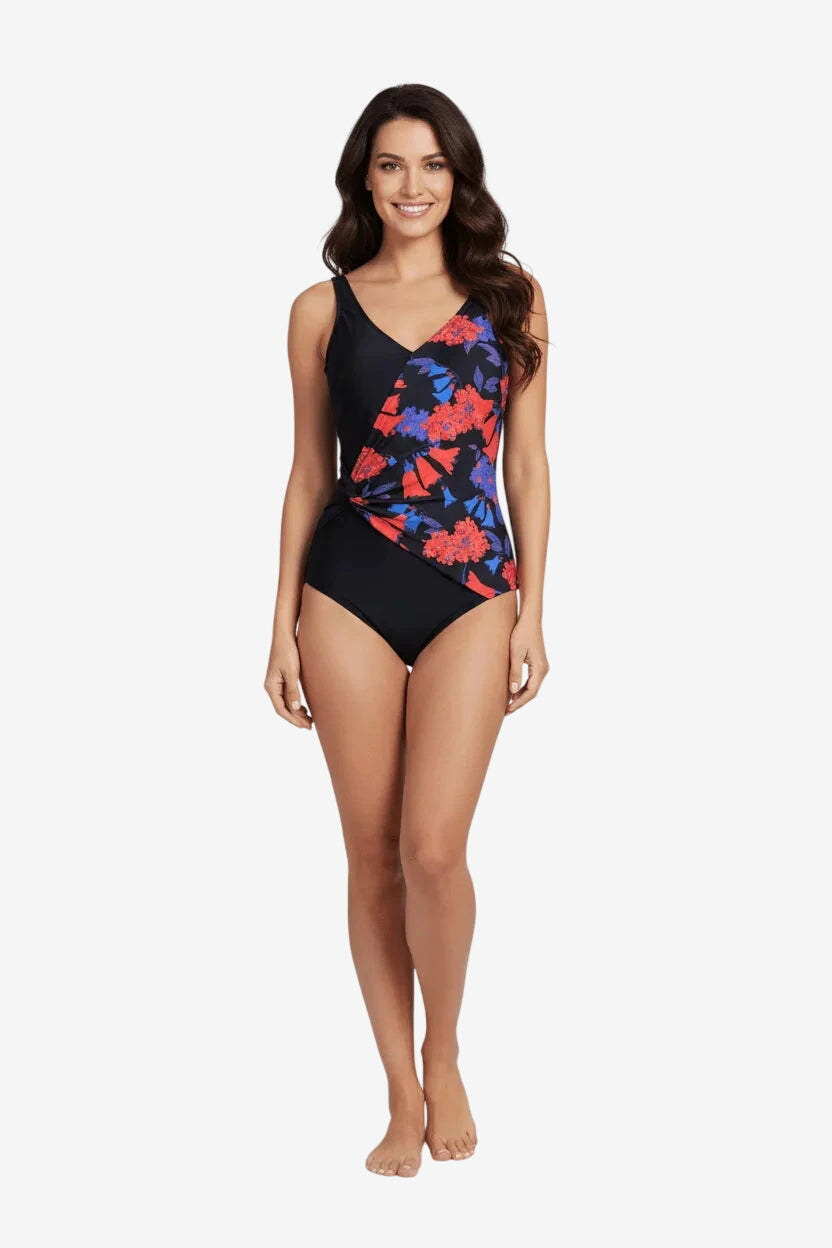 A woman wears a black one-piece swimsuit featuring a side-twist detail and a red and blue floral print.