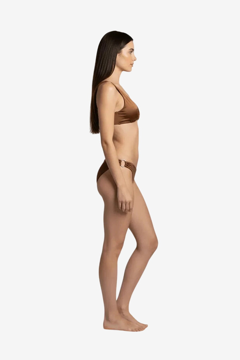 A woman stands in profile wearing a brown bikini with a gold ring detail on the hip.